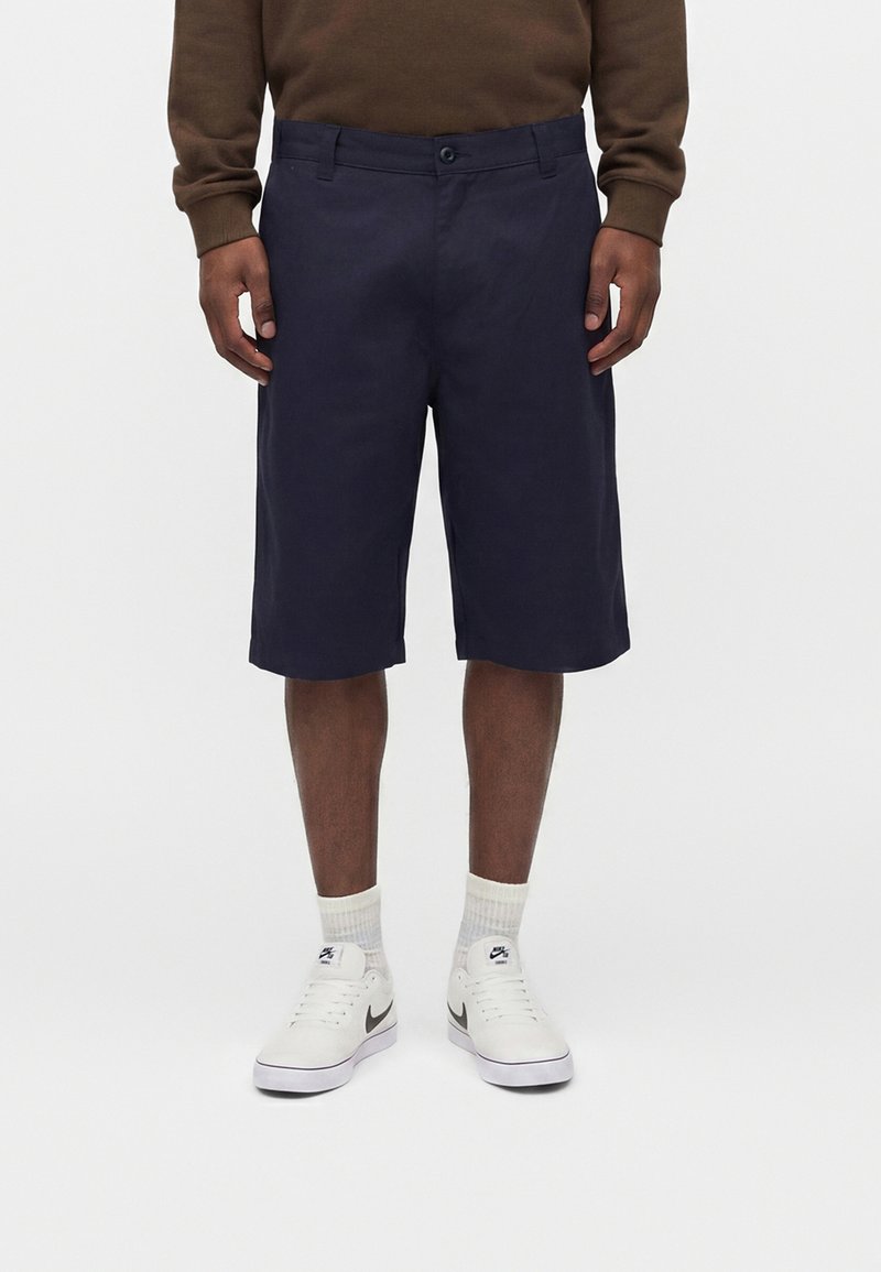 Mand iført marineblå knælange shorts, brun langærmet skjorte, hvide Nike-sneakers og hvide crew-sokker, der står mod en ensfarvet baggrund.