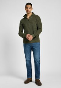 Khaki ribbed knit pullover dengan kerah ritsleting, dipadukan dengan celana jeans denim biru dan sepatu boots coklat, menampilkan desain yang pas dan kain yang bercorak.