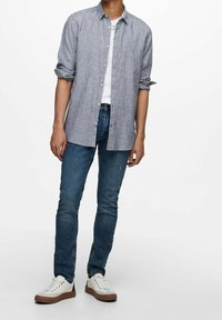 Camisa azul claro de botones con las mangas arremangadas, llevada sobre una camiseta blanca. Combinada con vaqueros ajustados azul y zapatillas blancas con suelas marrones.