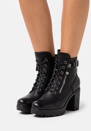 Mustat nahkaiset nilkkurit, joissa on nyöritys, paksut korot, sivuvetoketju ja solkikoriste, puettuna paljaisiin jalkoihin valkoista taustaa vasten.