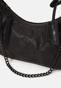 Sac à main en cuir noir avec motif de logo en relief, fermeture éclair et une bandoulière en chaîne épaisse pour le porter. Surface texturée avec un léger éclat.