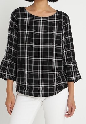 Czarna i biała kraciaste bluzka z długim rękawem i rozszerzanymi mankietami, noszona z białymi spodniami.