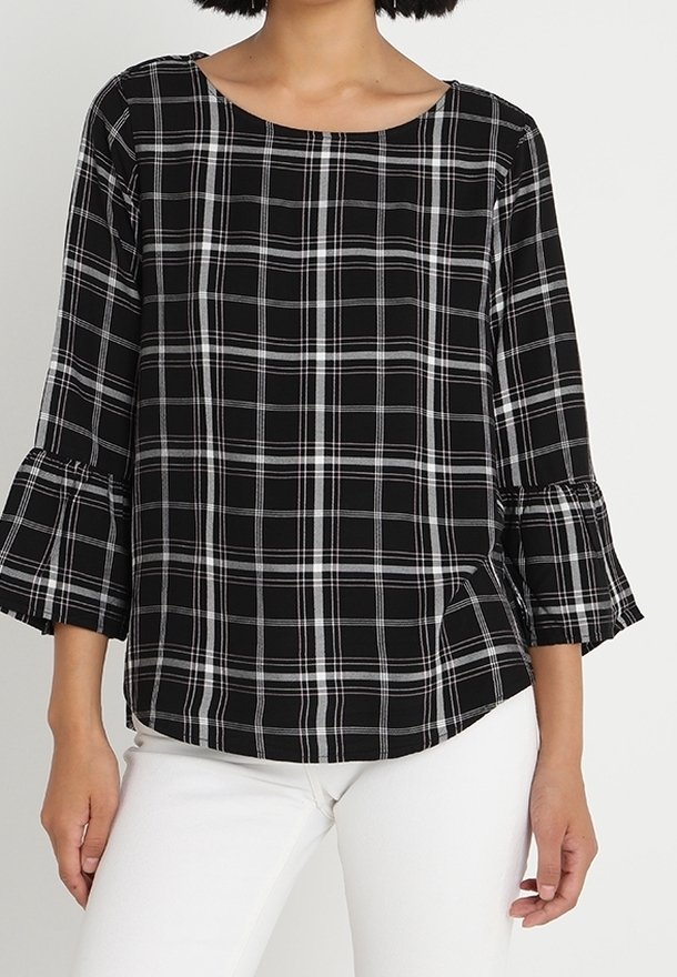 Blouse à manches longues en tissu à carreaux noir et blanc avec poignets évasés, portée avec un pantalon blanc.