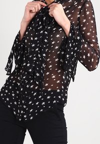 Svart genomskinlig blus med vitt stjärnmönster, volangärmar och knytbronsdetalj. Mjuk textur med en flödande design.