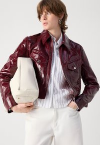 Burgundy, glossy leather jacket with a western design, pointed collar, chest pockets, and silver hardware, paired with a cream handbag.