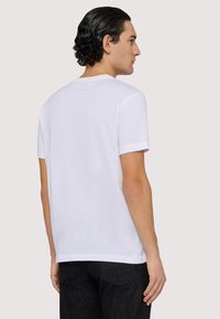 Biała bawełniana koszulka z krótkim rękawem, okrągłym dekoltem i luźnym krojem. Materiał jest gładki, z minimalistycznym wzornictwem.