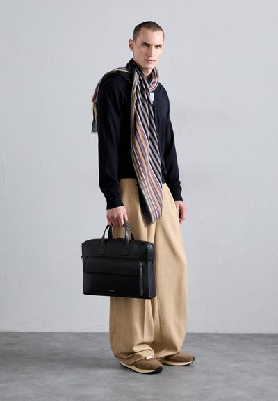 Black leather briefcase with dual handles, smooth texture, and front pocket. Model wears a navy sweater, striped scarf, and beige trousers.