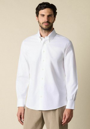 Man draagt een wit overhemd met lange mouwen en beige broek, staand tegen een effen beige achtergrond.