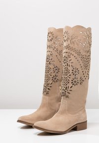 Botki w kolorze beżowym do kolan wykonane z zamszu, z wyciętymi wzorami kwiatowymi, falbaniastym wykończeniem na górze oraz niskim obcasem w kształcie bloku. Zamek błyskawiczny z boku umożliwia łatwe zakładanie.