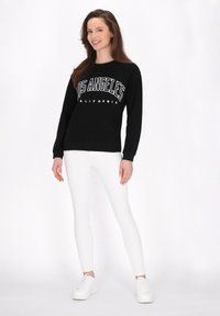 Sudadera negra con texto blanco "Los Ángeles California", combinada con pantalones ajustados blancos y zapatillas blancas, en un modelo contra un fondo claro.