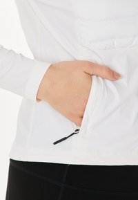 Vit långärmad topp med en dragkedjeficka och mjukt, lättviktigt tyg; inkluderar en svart dragrem och är tillsammans med svarta leggings.