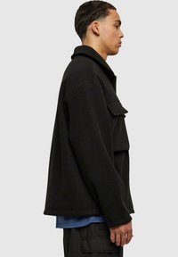 Schwarze Fleece-Jacke mit Kragen, Brusttasche und lockerer Passform. Über einem blauen Hemd getragen, Seitansicht angezeigt.