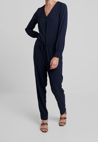 Combinaison bleu marine avec décolleté en V, manches longues, boutons sur le devant, cordon de serrage à la taille et jambes fuselées, associée à des sandales imprimé léopard.
