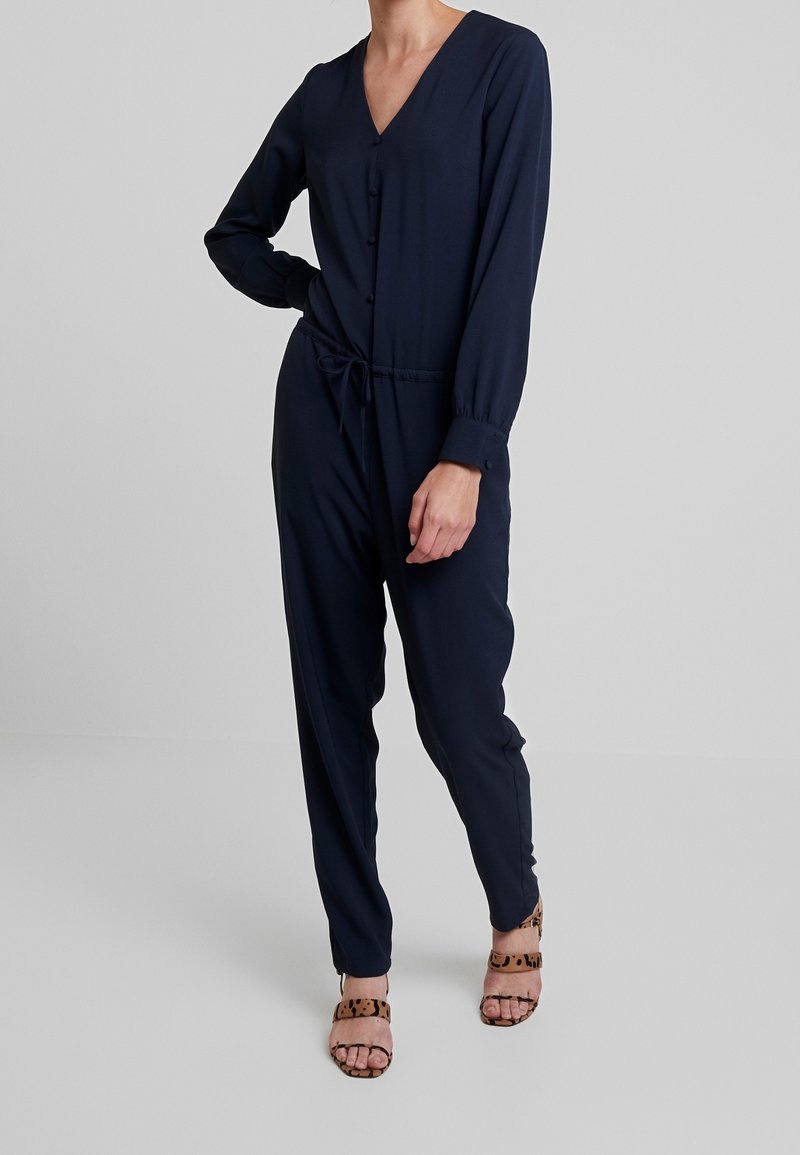 Combinaison bleu marine avec décolleté en V, manches longues, boutons sur le devant, cordon de serrage à la taille et jambes fuselées, associée à des sandales imprimé léopard.