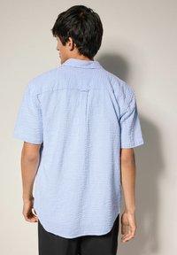 Camicia azzurra a maniche corte con un motivo a quadretti testurizzato, colletto con bottoni e orlo curvo. Spacchetto posteriore visibile e una piega singola sul retro.