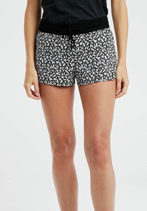 Mujer con pantalones cortos negros con pequeño estampado floral blanco y rosa y una camiseta negra, de pie con las manos relajadas a los lados.