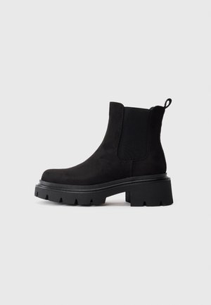 Schwarzer Wildleder-Stiefelette mit klobiger Profilsohle, elastischen Seiteneinsätzen und Zugschlaufe hinten vor hellem grauem Hintergrund.