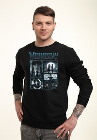 Mann in schwarzem Sweatshirt mit sechs bläulich getönten Szenen aus der TV-Show "Wednesday" und dem Titel in großer Schrift darüber.