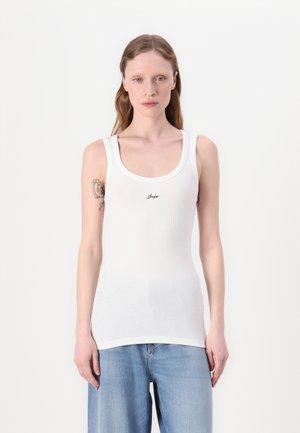 Weißes, geripptes Tanktop mit Rundhalsausschnitt, das auf der Vorderseite ein kleines schwarzes Logo trägt. Das Modell trägt es mit lässigen, lockeren blauen Jeans.