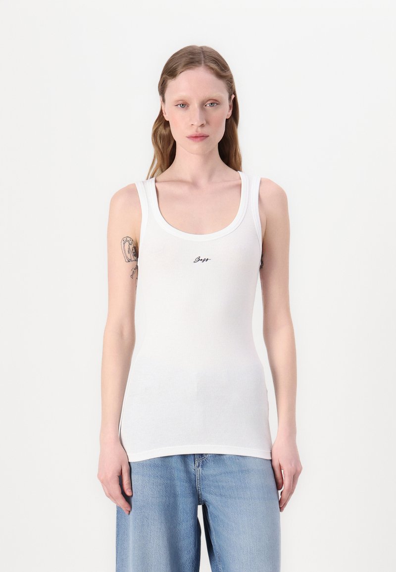 Camiseta sin mangas blanca con canalé y escote redondeado, que presenta un pequeño logo negro en la parte frontal. El modelo la lleva con unos vaqueros azules de corte holgado.