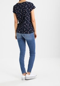 Kvinna som står med ryggen mot oss, iförd en marinblå kortärmad skjorta med rosa fågeltryck, ljusblå skinny jeans och vita platåskor.