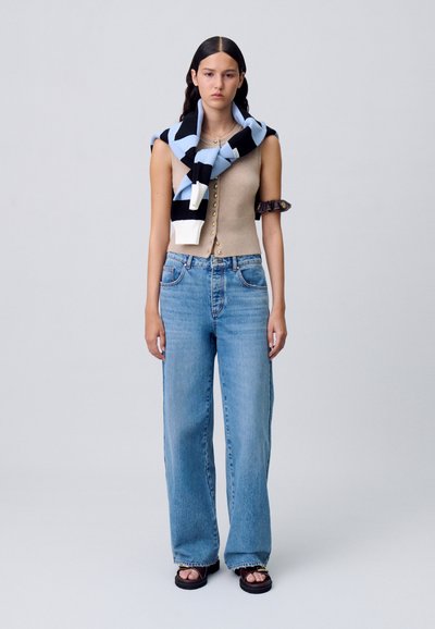 Top in maglia aderente beige con bottoni dorati, sciarpa a strisce blu chiaro e nera drappeggiata sulle spalle, jeans blu a gamba larga e sandali neri.