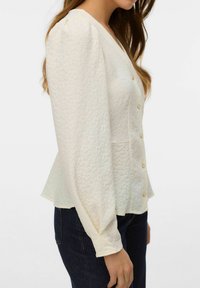 Jasnokremowa bluzka peplum z fakturowanej tkaniny, z długimi bufkami na rękawach, głębokim dekoltem w serek oraz ozdobnymi złotymi guzikami na przodzie.