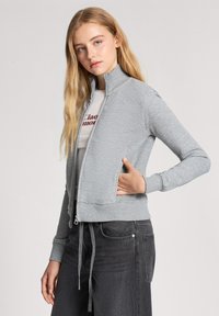 Felpa zip grigia chiara con colletto alto e polsini a coste, dotata di tasche laterali e design affusolato, abbinata a jeans scuri.