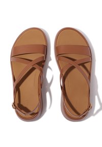 Bruine leren sandalen met dunne kruisbandjes, platte zool en een verstelbare enkelband met een goudkleurige gesp. Overal een gladde textuur.