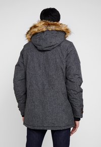 Chaqueta acolchada en gris carbón con capucha forrada de piel sintética, tejido texturizado y acentos de botones marrones en los puños. La vista trasera muestra las líneas de diseño.