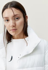 Chaqueta de plumas blanca con cuello alto, con textura acolchada y acabado brillante. Llevada sobre una camiseta blanca con estampado geométrico negro.