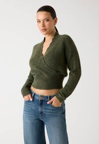 Pulover tricotat de culoare verde măsliniu, cu decolteu adânc în V, design scurt și textură striată. Este purtat cu blugi din denim albastru, cu talie înaltă.