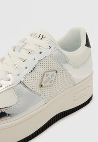 Baskets blanches avec des accents argentés brillants, des côtés perforés, des lacets plats et une semelle en caoutchouc noir épaisse. Présente un badge de logo sur le côté.