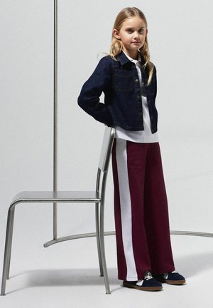 Chaqueta de mezclilla con botones metálicos, camisa blanca y pantalones de pierna ancha en color burdeos con una franja blanca en el costado. Zapatos negros con cordones blancos.