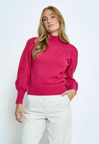 Aangepaste roze trui met hoge nek, gestructureerde gevlochten accenten op de mouwen en ribbelzoom. Gecombineerd met lichte broek.