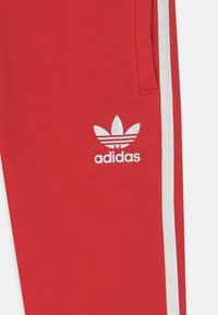 Červené tepláky s bílými bočními pruhy a bílým logem Adidas. Vyrobeny z texturované, měkké látky s volným střihem.
