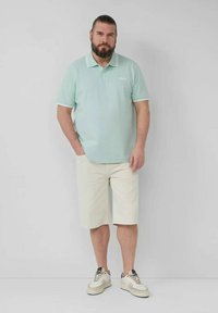 Helles grünes Poloshirt mit weißen Akzenten, kombiniert mit beigen Shorts und Sneakers. Das Model steht mit den Händen in den Taschen vor einem neutralen Hintergrund.