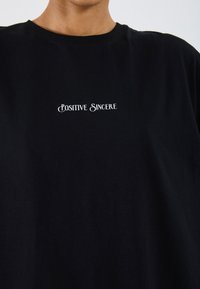 Schwarzes Baumwoll-T-Shirt mit "POSITIVE SINCERE" in weißen, gestickten Buchstaben zentral auf der Brust, mit Rundhalsausschnitt.
