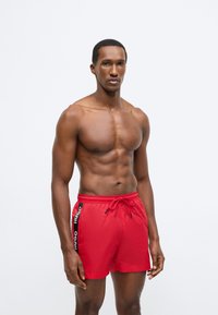 Piros, könnyű anyagból készült fürdőnadrág, fekete oldalsávval, melyen "HUGO" felirat található, rugalmas derékpánttal és húzózsinóros rögzítéssel.