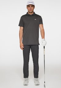 Mörkgrå pikétröja med krage, matchande mörkgrå byxor. Bär en vit keps och håller en golfklubba i en handske.