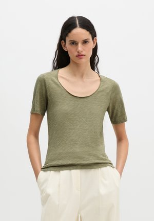 Jeune femme aux longs cheveux foncés portant un t-shirt vert olive à manches courtes et encolure ronde, ainsi qu'un pantalon blanc, debout, les mains dans les poches.