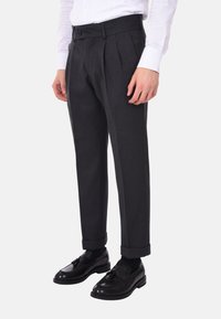 Pantaloni grigi a pieghe con una vestibilità sartoriale, realizzati in un tessuto liscio. Presentano una chiusura con bottone e orli arrotolati, abbinati a scarpe nere.