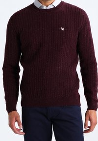 Viininpunainen neulottu neule, jossa on ribbineulosta, pyöreä kaulus ja pieni kirjailtu logo rinnassa. Kangas sisältää hienovaraisia pilkkuja.