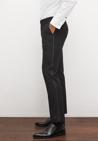 Muž nosi čierne spoločenské nohavice s bočným prúžkom, bielu košeľu a čierne spoločenské topánky, stojí bokom s jednou rukou v vrecku.