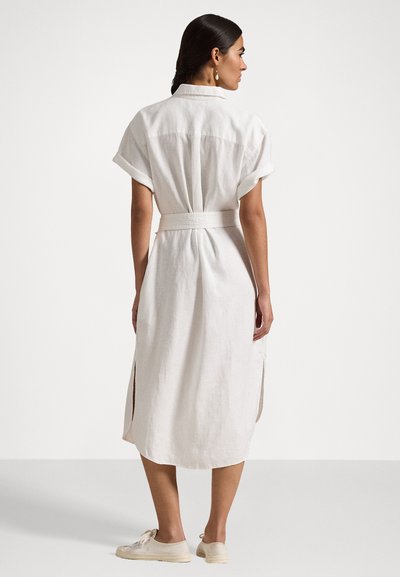 Robe chemise blanche en lin avec des manches courtes, taille ceinturée, fentes sur les côtés et ourlet arrondi, associée à des baskets de couleur claire.