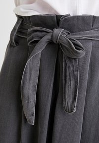 Gros plan sur un pantalon gris foncé taille haute avec ceinture en tissu nouée en un nœud à la taille, porté avec un haut blanc rentrée à l'intérieur.