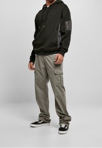 Hombre con sudadera negra con bolsillo con cremallera en la manga, pantalones cargo gris claro y zapatillas negras con suelas blancas, de pie sobre un fondo gris.