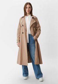 Beige trenchcoat met dubbelzijdige knopen, brede revers en een relaxte pasvorm, gecombineerd met lichtblauwe flared jeans en witte sneakers.
