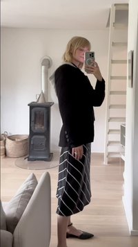 Črn blazer z gumbi in kapuco, v kombinaciji s midi krilom v črno-beših črtah. Model nosi ravne čevlje in stoji v modernem interierju.