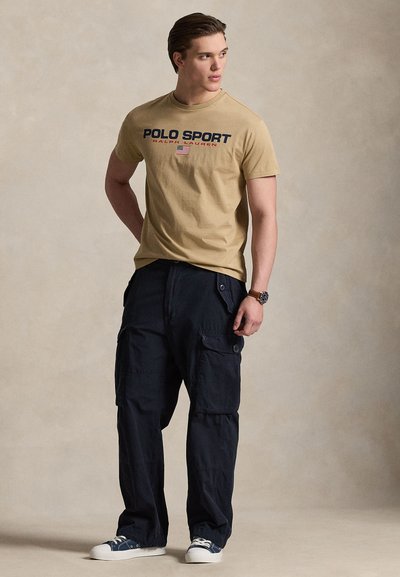 Μπεζ βαμβακερό t-shirt με το λογότυπο "POLO SPORT", σε συνδυασμό με σκούρο μπλε cargo παντελόνι με πλαϊνές τσέπες και κουμπί, φορεμένο με σκούρα μπλε αθλητικά παπούτσια.
